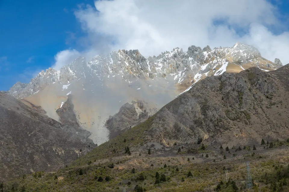 蓝天和云雾之间的锯齿状雪山山脊，前景是零星树木覆盖的山坡。